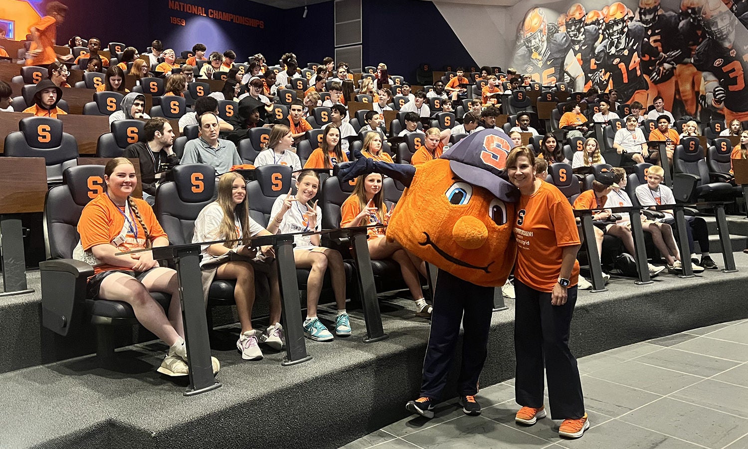A large audience in orange attire sits in an auditorium with Syracuse University-branded seats. Syracuse’s “Otto the Orange” mascot and an event organizer are visible at the front.